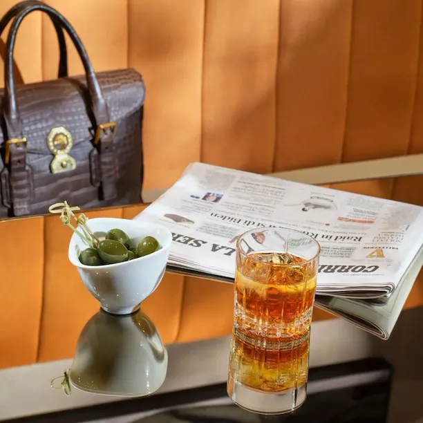 Close up on one of the tables in Il Salotto Lobby Lounge with a cocktail, green olives and a newspaper. Leather work bag in the background