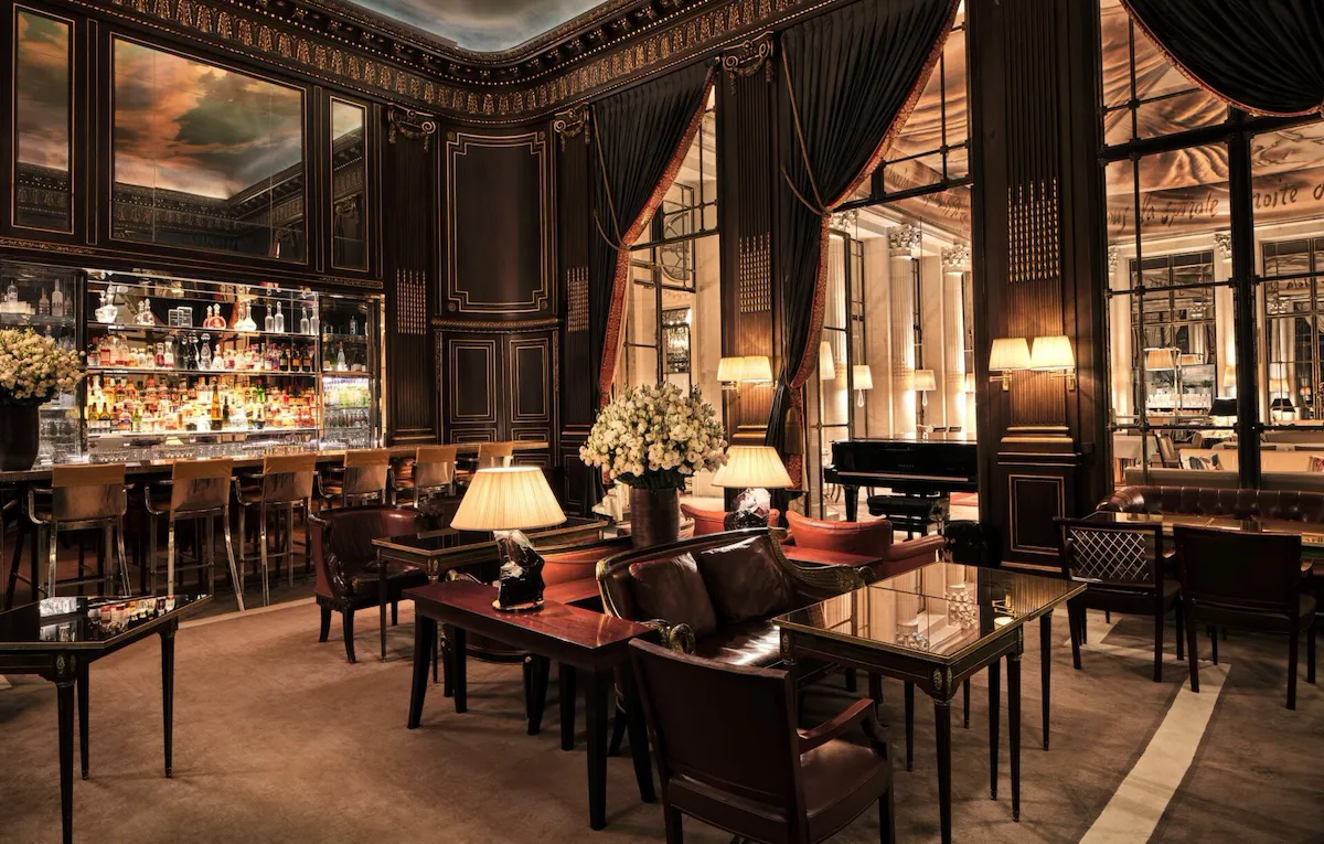 Au centre de la photo, on voit des lampes allumées sur des tables avec des fauteuils et des coussins rouge foncé, et un bouquet de fleurs blanches dans un vase. À gauche, le bar 228 avec des chaises hautes. On peut voir les bouteilles sur les étagères. Au-dessus du bar, un miroir reflète le plafond, qui représente un ciel avec des nuages. Nous regardons les boiseries sur les murs, avec ses détails dorés. À droite, un piano et de grandes fenêtres avec des rideaux rouges, révélant une autre pièce du Meurice, Paris.