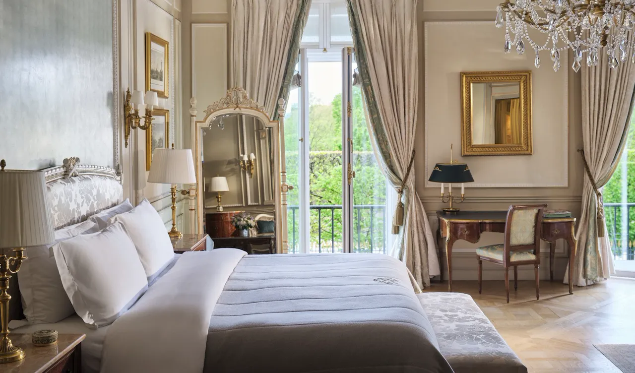 Large view of the right side of the bed with the mirror, the desk, the chair and the windows open behind, at Le Meurice, Paris