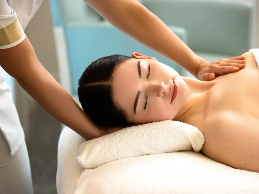 We can see a woman lying on a massage table, resting her head on a white pillow. We can see the client's face positioned on her side. She has her eyes closed and her hair is tied back. On the left-hand side of the photo, we can see the masseur's uniform. One of the hands is positioned under the client's head and the second hand is on her upper chest. We can see the white blanket covering the client's chest at Le Meurice, Paris.